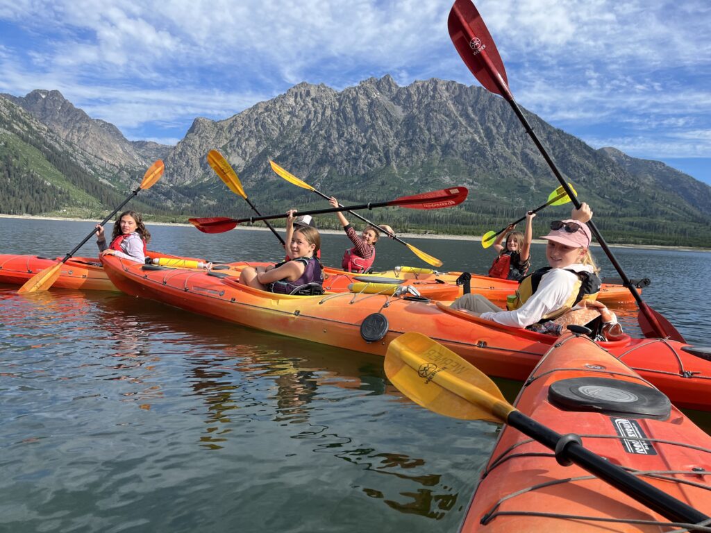 Image of GAP! girls out on the lake in kayaks with paddles raised over their heads.